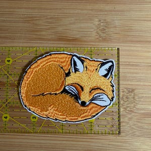 May include: An embroidered patch of a curled-up orange fox with white accents and black details. The patch is positioned on a yellow ruler, showing it measures approximately 3 inches in width and 2 inches in height.