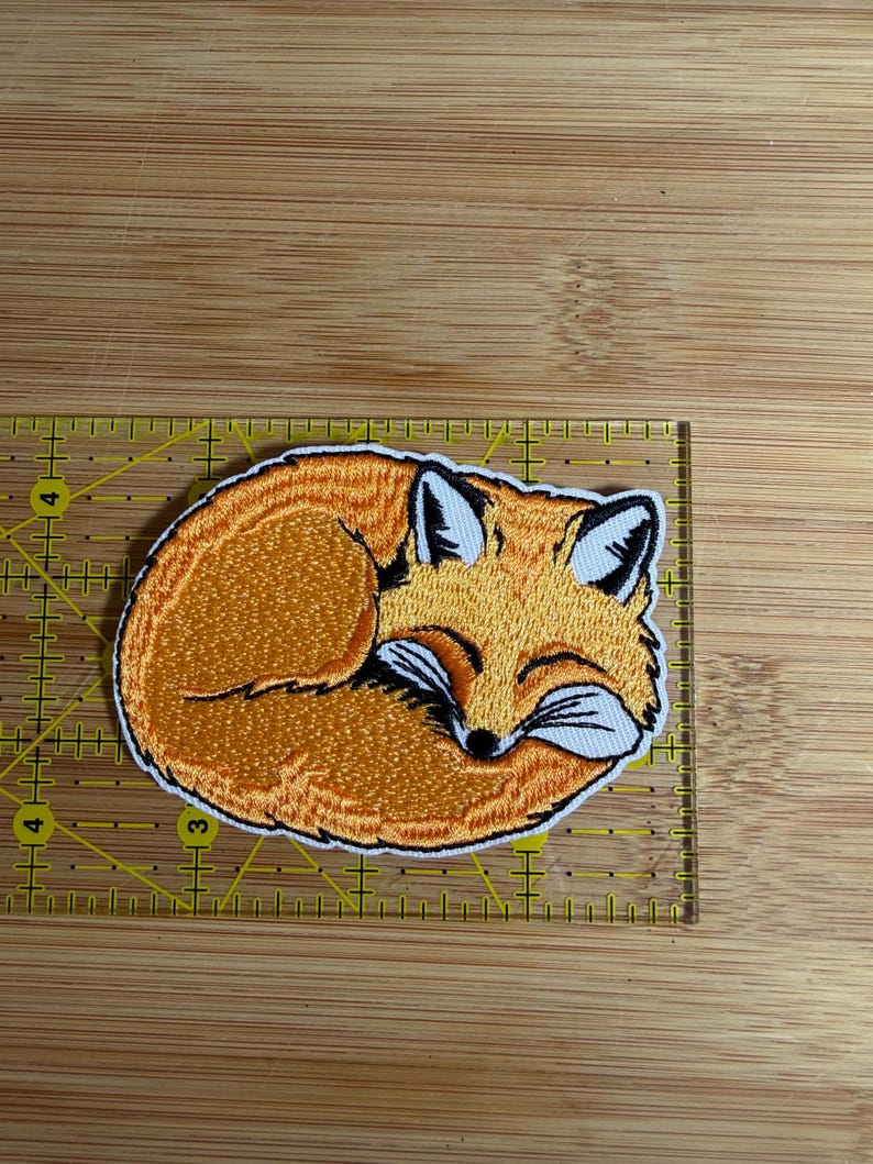 Op de afbeelding: Een geborduurde patch van een opgerolde oranje vos met witte accenten en zwarte details. De patch ligt op een gele liniaal en meet ongeveer 7,5 cm in de breedte en 5 cm in de hoogte.