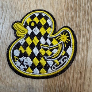 May include: An embroidered patch shaped like a rubber duck featuring a black, yellow, and white checkered pattern. The duck has a glittery black border, a yellow beak, and includes small embroidered icons of a sun, a crescent moon, and a star.