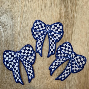 May include: Three embroidered patches in the shape of bows, each featuring a blue and white checkered pattern. The patches have a distinct blue border outlining the bow shape, with two loops and two trailing ribbon ends on each piece.