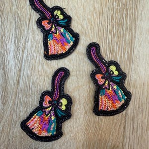 May include: Three embroidered patches shaped like brooms with black borders. Each patch features a handle, a bow, and bristles stitched in vibrant multicolored threads including orange, purple, yellow, and teal against a black background.