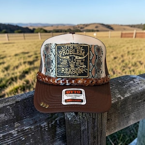 Peut inclure: Une casquette trucker marron avec un panneau avant crème à motif peau de serpent et un écusson rectangulaire noir. L'écusson présente un crâne de taureau et le texte IF I'M THE PROBLEM THEN YOU MIGHT BE THE REASON. Une chaîne marron orne la visière.