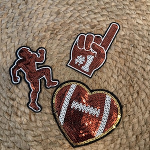 Puede incluir: Tres parches decorativos que incluyen un jugador de fútbol americano de lentejuelas marrones en pose de carrera, un dedo de espuma de lentejuelas marrones con el texto #1 y un balón de fútbol en forma de corazón con lentejuelas y cordones blancos.