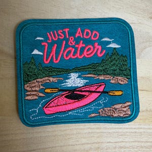 May include: A square embroidered patch with a teal background featuring a pink kayak on water, brown rocky shores, green pine trees, and white clouds. The text "JUST ADD & Water" is stitched in pink above the kayak, with a yellow and black paddle.