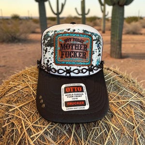 May include: A black and white trucker hat featuring a foam front panel with a distressed cow print pattern. A rectangular leather patch displays the text NOT TODAY MOTHER FUCKER. A black barbed wire trim accents the base of the front panel.