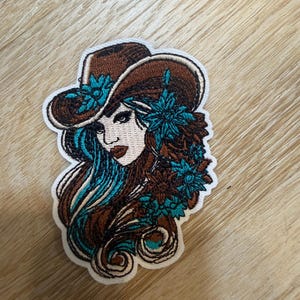 May include: An embroidered patch featuring a person wearing a brown cowboy hat adorned with teal flowers. The person has long brown hair with teal highlights and is decorated with additional teal floral accents on the side of their hair.