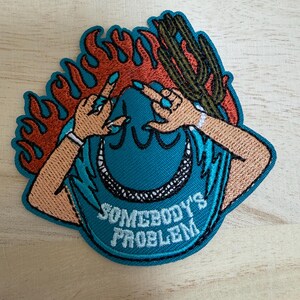 Puede incluir: Un parche bordado que muestra un sombrero de vaquero azul con el texto SOMEBODY'S PROBLEM, rodeado de llamas naranjas y un cactus verde. El diseño incluye dos manos con uñas azules posicionadas sobre el sombrero, todo bordeado en azul.