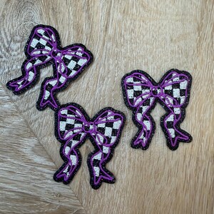 May include: Three embroidered patches shaped like bows, each featuring a black and white checkered pattern. The bows are outlined in vibrant purple thread and have a subtle glittery texture throughout the black and white fabric sections.