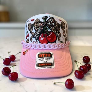 Puede incluir: Una gorra de camionero blanca y rosa con un gráfico central de dos cerezas rojas atadas con un lazo de estampado de leopardo. La gorra incluye una cadena de plástico rosa claro sobre la visera, un pequeño dije de cereza y una etiqueta que dice OTTO WORLD FAMOUS FOAM FRONT.