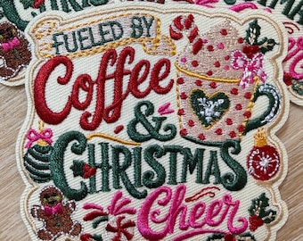 Może przedstawiać: Prostokątna haftowana naszywka na kremowym tle z napisem FUELED BY Coffee & CHRISTMAS Cheer. Wzór zawiera kubek w kropki z laską cukrową, piernikowego ludzika z różową kokardką oraz świąteczne ozdoby bożonarodzeniowe.