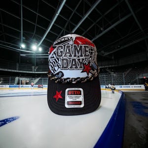 May include: A white and black trucker hat featuring a glittery black text graphic that reads GAME DAY, a red star, and a hockey-themed design. The hat includes a thick black chain across the brim and a rectangular OTTO brand sticker on the front.