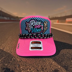 May include: A bright pink trucker hat featuring a blue and black checkered pattern on the front panel. An embroidered patch reads Race Day Vibes with star and lightning bolt accents. A black chain with rhinestones sits above the brim.