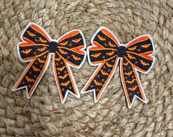 Könnte beinhalten: Zwei gestickte Schleifen-Aufnäher mit schwarzem Hintergrund und einem wiederkehrenden Muster aus kleinen orangefarbenen Fledermäusen und Sternen. Jede Schleife hat einen schwarzen Mittelknoten und einen dicken weißen Rand.
