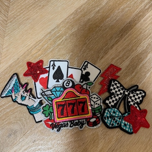 May include: A collection of embroidered patches featuring Las Vegas themes, including playing cards, a slot machine with the text Vegas Baby, dice, cherries, a martini glass, stars, and a lightning bolt in red, black, white, blue, and silver glitter.