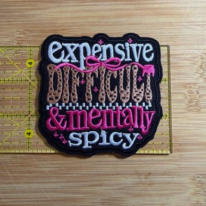 Op de afbeelding: Een zwarte geborduurde patch met de tekst 'expensive DIFFICULT & mentally spicy' in wit, luipaardprint en felroze garen, versierd met een roze strik en kleine sterretjes, met een breedte van ongeveer 9 cm.