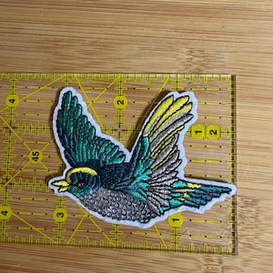May include: An embroidered bird patch featuring teal, yellow, and silver thread with a sequined belly. The patch is positioned on a yellow quilting ruler, showing it measures approximately 3.5 inches in width and 2.5 inches in height.