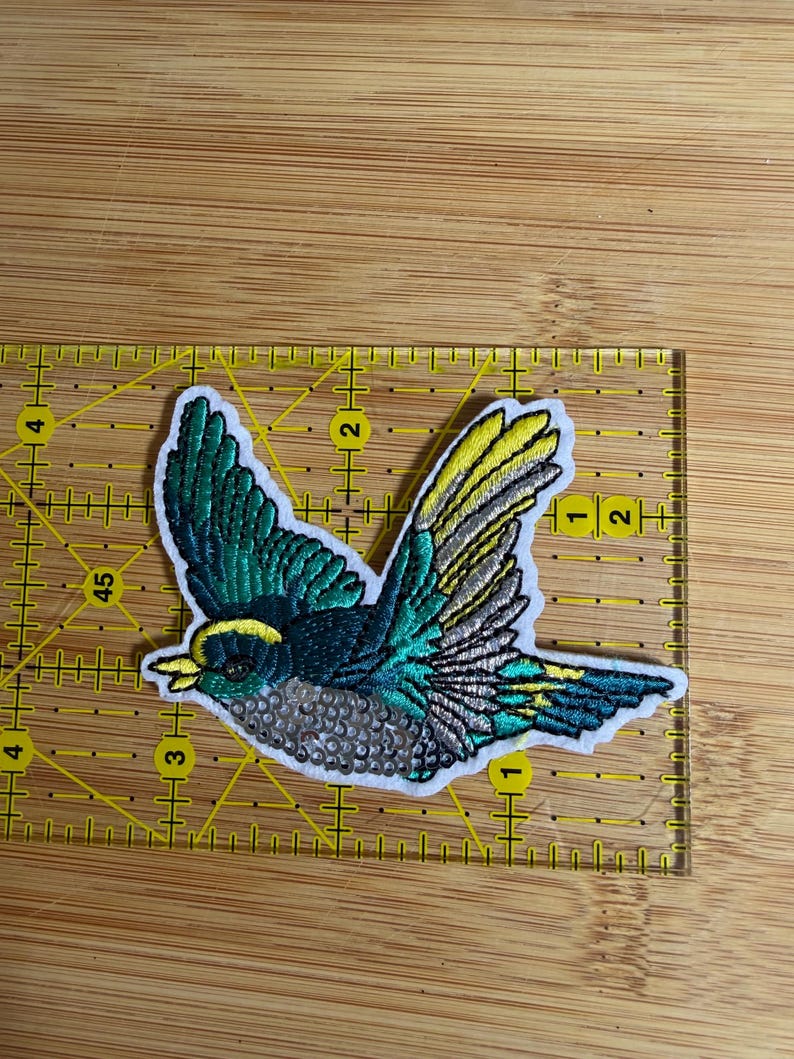May include: An embroidered bird patch featuring teal, yellow, and silver thread with a sequined belly. The patch is positioned on a yellow quilting ruler, showing it measures approximately 3.5 inches in width and 2.5 inches in height.