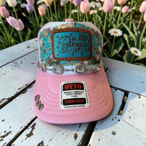 May include: A pink trucker hat with a white mesh back and a front panel featuring a floral pattern in shades of blue and brown. A rectangular patch with the text 'a lil' Bougie' is centred, accented by a rhinestone chain and three small crystals.