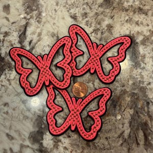 Pode incluir: Três emblemas bordados de borboleta com fio rosa néon vibrante e uma borda preta. Os emblemas estão dispostos em grupo com uma moeda de cobre colocada no centro para fornecer uma referência de escala sobre o tamanho dos objetos.