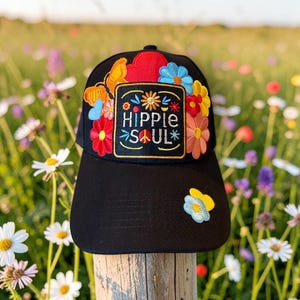 Puede incluir: Una gorra de béisbol negra con un parche rectangular central que presenta el texto bordado "HIPPIE SOUL" rodeado de motivos florales y de mariposas coloridos. Flores bordadas adicionales se encuentran en la copa y la visera.