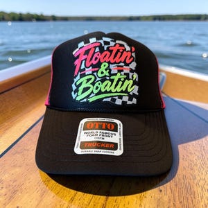 Puede incluir: Una gorra de camionero negra con frente de espuma que presenta un patrón de cuadros y el texto "Floatin' & Boatin'" en rosa y verde. La gorra incluye una pegatina blanca en la visera con el texto "OTTO WORLD FAMOUS FOAM FRONT OSFM TRUCKER".