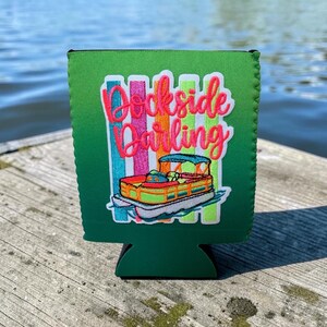 May include: A green foam beverage insulator featuring a colorful embroidered graphic of a pontoon boat. The text "Dockside Darling" is stitched in bright pink script above the boat, set against a background of vertical stripes in blue, pink, and yellow.