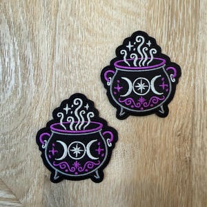 Puede incluir: Dos parches bordados que muestran un caldero negro con detalles en hilo morado y blanco. El diseño incluye un símbolo de triple luna, una estrella central, vapor en espiral y pequeños acentos de estrellas. Los parches tienen un borde negro y tienen forma de olla de tres patas con asas laterales.