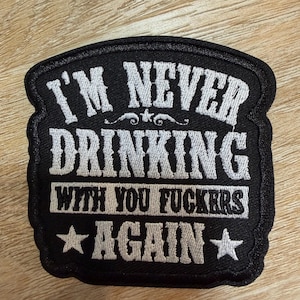 May include: A black embroidered patch with a white border and white text that reads I'M NEVER DRINKING WITH YOU FUCKERS AGAIN, featuring a small star above the word DRINKING and two stars flanking the word AGAIN on the bottom line.