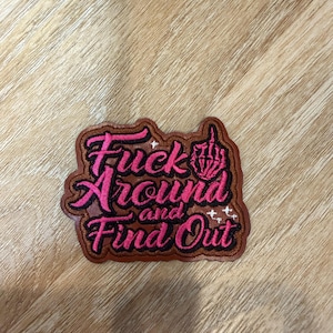 May include: A brown leather patch with embroidered pink text that reads "Fuck Around and Find Out." The design features a skeleton hand making a middle finger gesture, accented with small white star-like sparkles stitched into the leather.