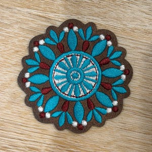 May include: A circular brown patch featuring intricate embroidery in bright turquoise, deep red, and white thread. The design consists of a central wheel-like pattern surrounded by radiating petal shapes and small dots, creating a symmetrical floral-inspired motif on a textured brown base.