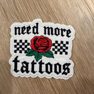 May include: A white embroidered patch with a scalloped edge featuring the black text "need more tattoos" in a gothic font. The design includes a central red rose with green leaves, flanked by two black and white checkered square patterns.