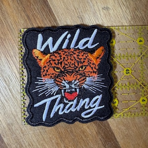 Puede incluir: Un parche bordado rectangular con fondo negro que presenta una cabeza de leopardo naranja y negra con bigotes blancos y una lengua roja. El texto "Wild Thang" está bordado en letra blanca sobre y debajo del leopardo. Una regla amarilla es visible a lo largo del borde derecho, mostrando que el parche mide aproximadamente 7,6 cm de ancho.