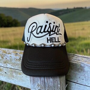 May include: A black and white trucker hat featuring the text Raisin' HELL in black embroidery on the white front panel. The hat includes a decorative silver-toned chain trim with small white oval accents across the base of the front panel.