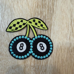 May include: An embroidered patch shaped like two cherries, where each fruit is a black and white 8-ball surrounded by a border of bright blue stitching. The two stems connect to a pair of lime green leaves featuring a black checkered pattern.