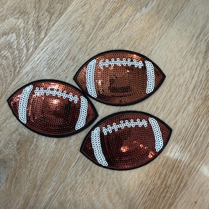 Puede incluir: Tres parches con forma de balón de fútbol americano cubiertos de lentejuelas marrones. Cada parche presenta un borde negro y detalles de costura en lentejuelas blancas que imitan los cordones y las rayas de un balón de fútbol americano.