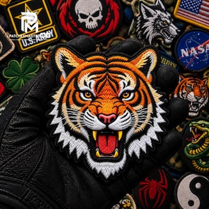Könnte beinhalten: Ein gestickter Aufnäher in Form eines Tigerkopfes mit detaillierten orangefarbenen, schwarzen und weißen Fäden. Der Tiger ist mit geöffnetem Maul, entblößten gelben Fangzähnen und roter Zunge dargestellt, vor einem Hintergrund aus weiteren Aufnähern.