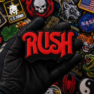 May include: An embroidered patch featuring the word RUSH in bold red lettering with a black border. The design has a distinct stylized font with sharp edges. The patch is held against a background of various other embroidered patches.