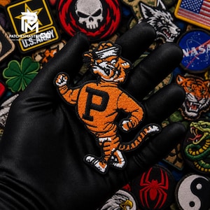 May include: An embroidered patch featuring an orange tiger with black stripes, wearing a white sailor hat, and displaying a large black letter P on its chest. The tiger is depicted in a dynamic pose with one arm raised and a curved tail.