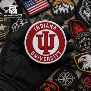 Puede incluir: Un parche bordado circular que presenta el logotipo de la Universidad de Indiana en rojo y blanco. El diseño incluye las letras IU en el centro con el texto INDIANA UNIVERSITY arqueado alrededor del borde. El parche se sostiene sobre un fondo de varios otros parches bordados.