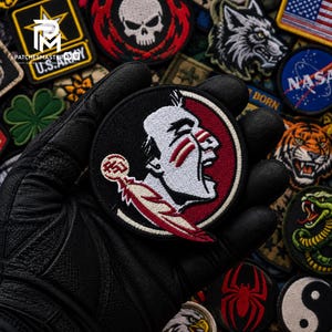 May include: A circular embroidered patch featuring a profile of a person with white skin, red face paint, and a red and white feather. The design includes a maroon background and a small circular emblem with the letters FSU in white and red.