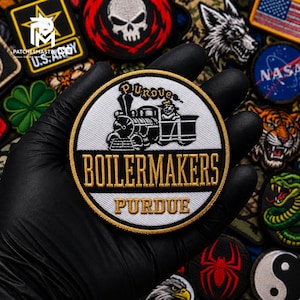 Op de afbeelding: Een ronde geborduurde patch met een zwart-witte stoomlocomotiefafbeelding boven de tekst BOILERMAKERS en PURDUE in gouden letters. Het ontwerp is geplaatst tegen een witte achtergrond met een dikke gouden rand en zwarte afwerking.