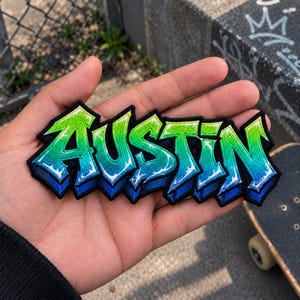 May include: An embroidered patch featuring the word AUSTIN in a graffiti-style font. The letters transition from a bright lime green at the top to a deep blue at the bottom, with a thick black border outlining the entire shape of the text.