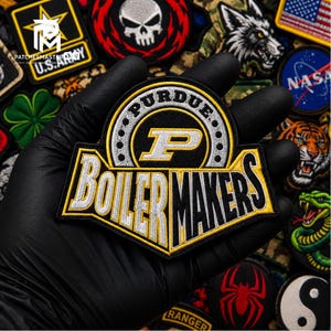 Puede incluir: Un parche bordado en negro, dorado y blanco con el texto Purdue Boilermakers y una letra P grande. El parche tiene una sección superior circular con un borde de puntos pequeños y una base más ancha con letras en negrita y bordes cosidos.
