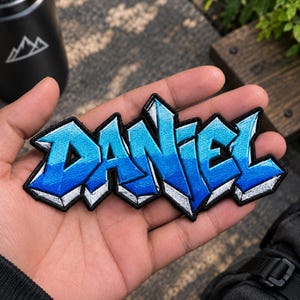 May include: A fabric patch featuring the name DANIEL in a stylized, blocky graffiti font. The letters are colored in a gradient of light blue to dark blue, outlined in black with white highlights to create a three-dimensional effect.