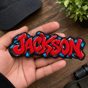 Puede incluir: Un parche bordado rectangular que muestra el nombre JACKSON en letras rojas estilo grafiti con un contorno negro y un efecto de sombra azul brillante. El parche tiene una superficie de tela texturizada y está diseñado para fijarse en ropa o equipo.