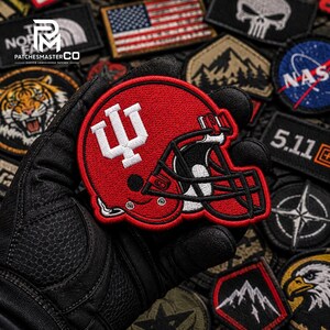 Peut inclure: Un écusson brodé en forme de casque de football américain rouge avec un logo IU blanc sur le côté et une grille de protection noire. L'écusson présente des coutures détaillées et est présenté sur un fond composé d'autres écussons brodés.