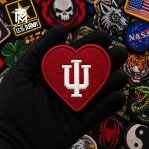 Peut inclure: Un écusson brodé en forme de cœur avec un fond rouge profond et le logo blanc IU de l'Université de l'Indiana au centre. L'écusson présente une bordure rouge épaisse et est présenté devant un assortiment d'autres écussons brodés variés.