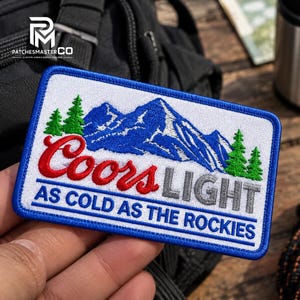 May include: A rectangular embroidered patch with a white background and a blue border. It features blue mountain peaks, green pine trees, and the red text Coors, the grey text LIGHT, and the blue text AS COLD AS THE ROCKIES.