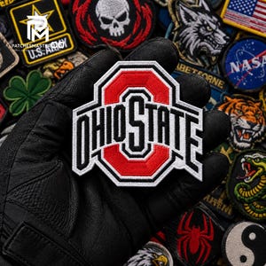 May include: An embroidered Ohio State University patch featuring a large red block O with the word OHIO STATE in black lettering, outlined in white, centered on a black background. The patch is held against a collection of various other patches.