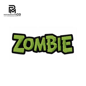 Könnte beinhalten: Ein gestickter Aufnäher mit dem Wort ZOMBIE in leuchtend hellgrünen Blockbuchstaben mit einem dicken schwarzen Rand. Der Text ist in einer zackigen Cartoon-Schriftart gestaltet und vor einem weißen Hintergrund abgebildet.